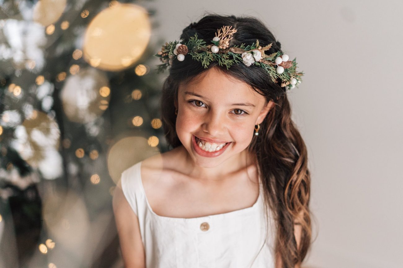 Girl in a white dress and festive crown grinning, Christmas tree lights glowing behind her