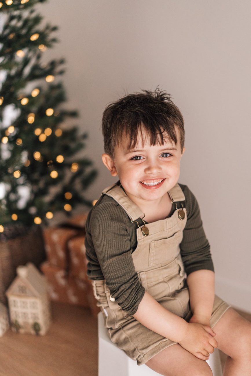 Little boy in overalls laughing on a stool beside the Christmas tree