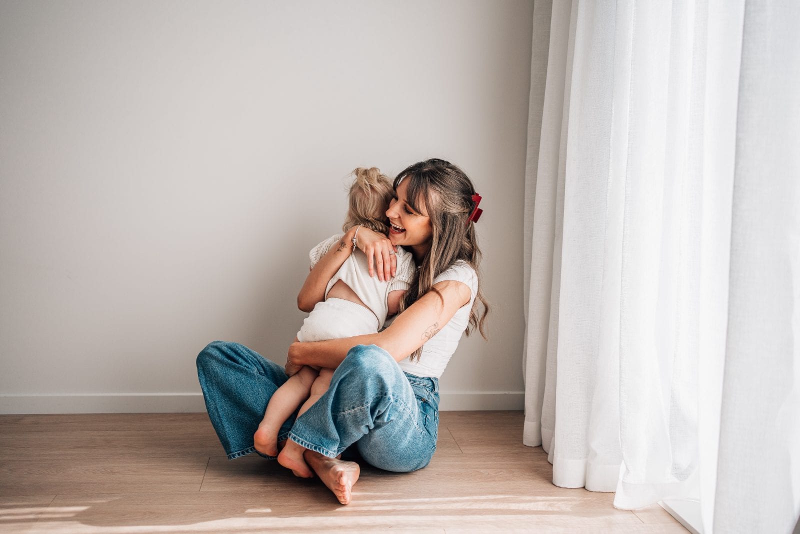 Mother in jeans sitting on the studio floor cuddling her toddler daughter beside a sheer white curtain.
