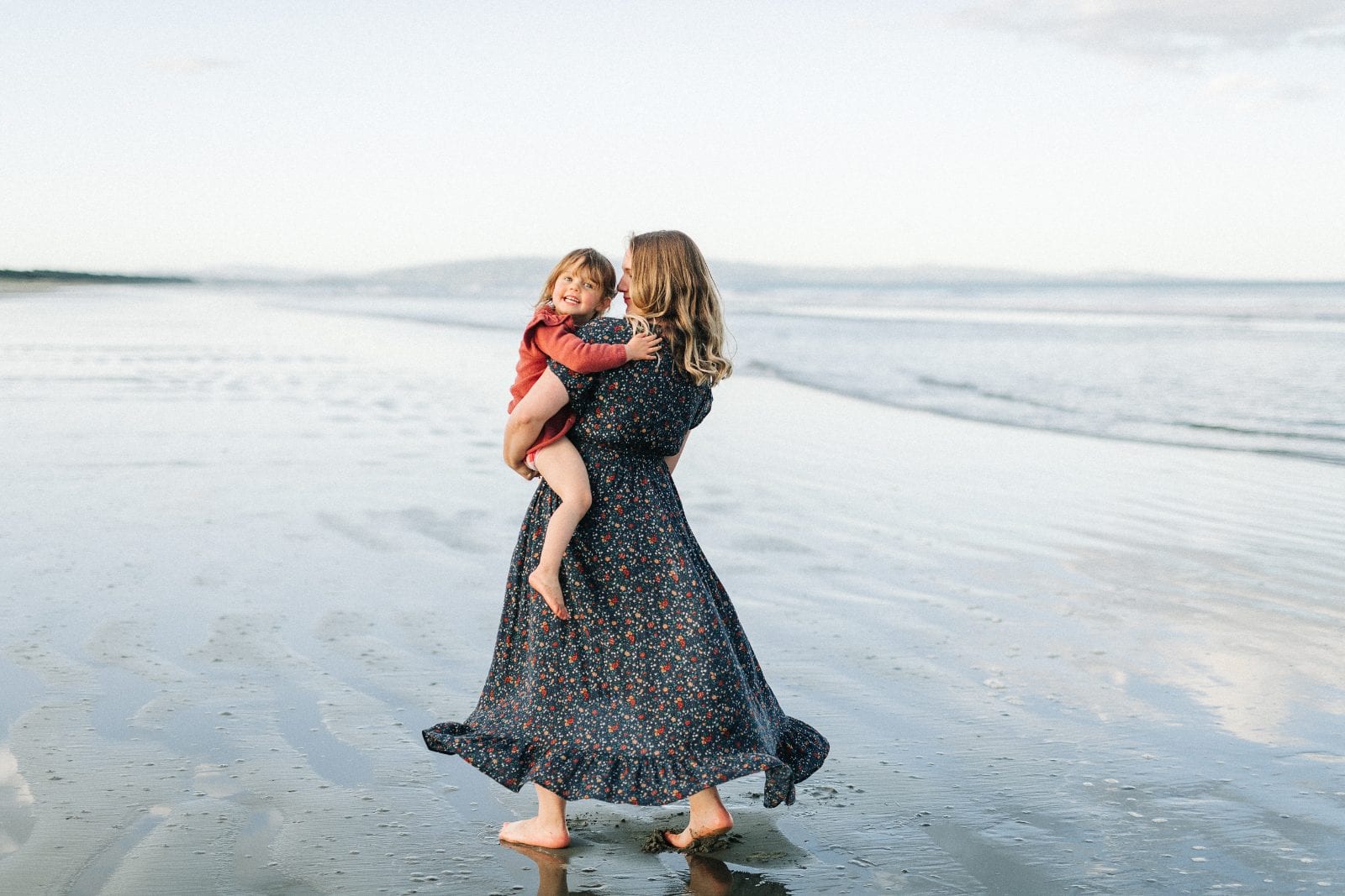 A mother in a floral dress holds her young daughter on a wide sandy beach at golden hour in Canterbury.