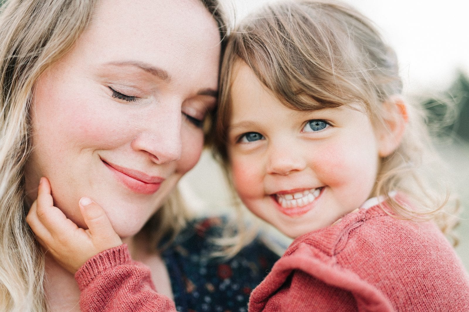 A mother and her bright-eyed young daughter smile cheek to cheek in soft natural light.