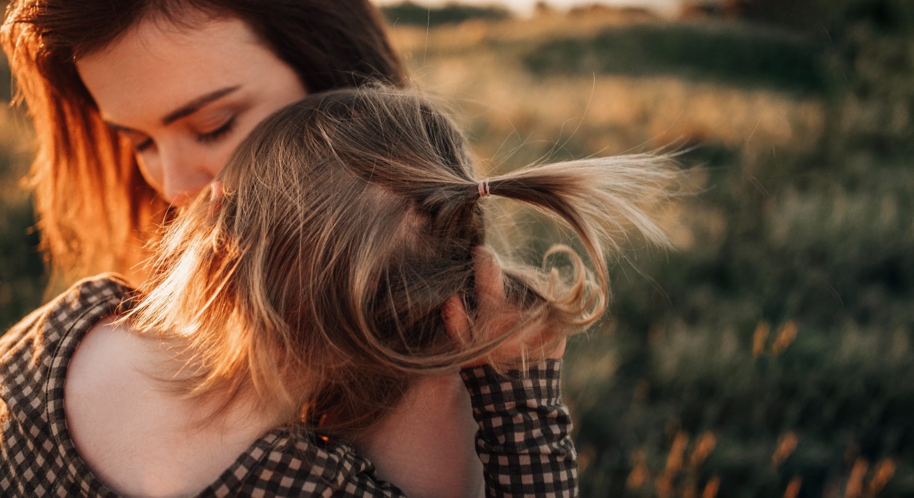 A mother nuzzles her young daughter's hair in a grassy field bathed in warm golden-hour backlight.