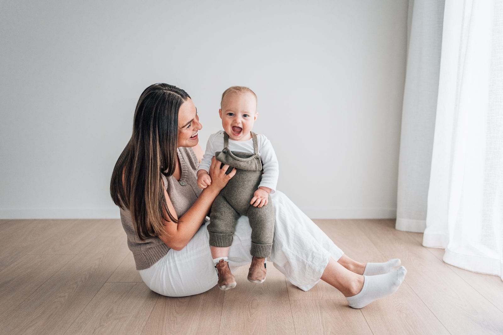 Mother sitting on the studio floor holding her laughing baby boy in knitted overalls.