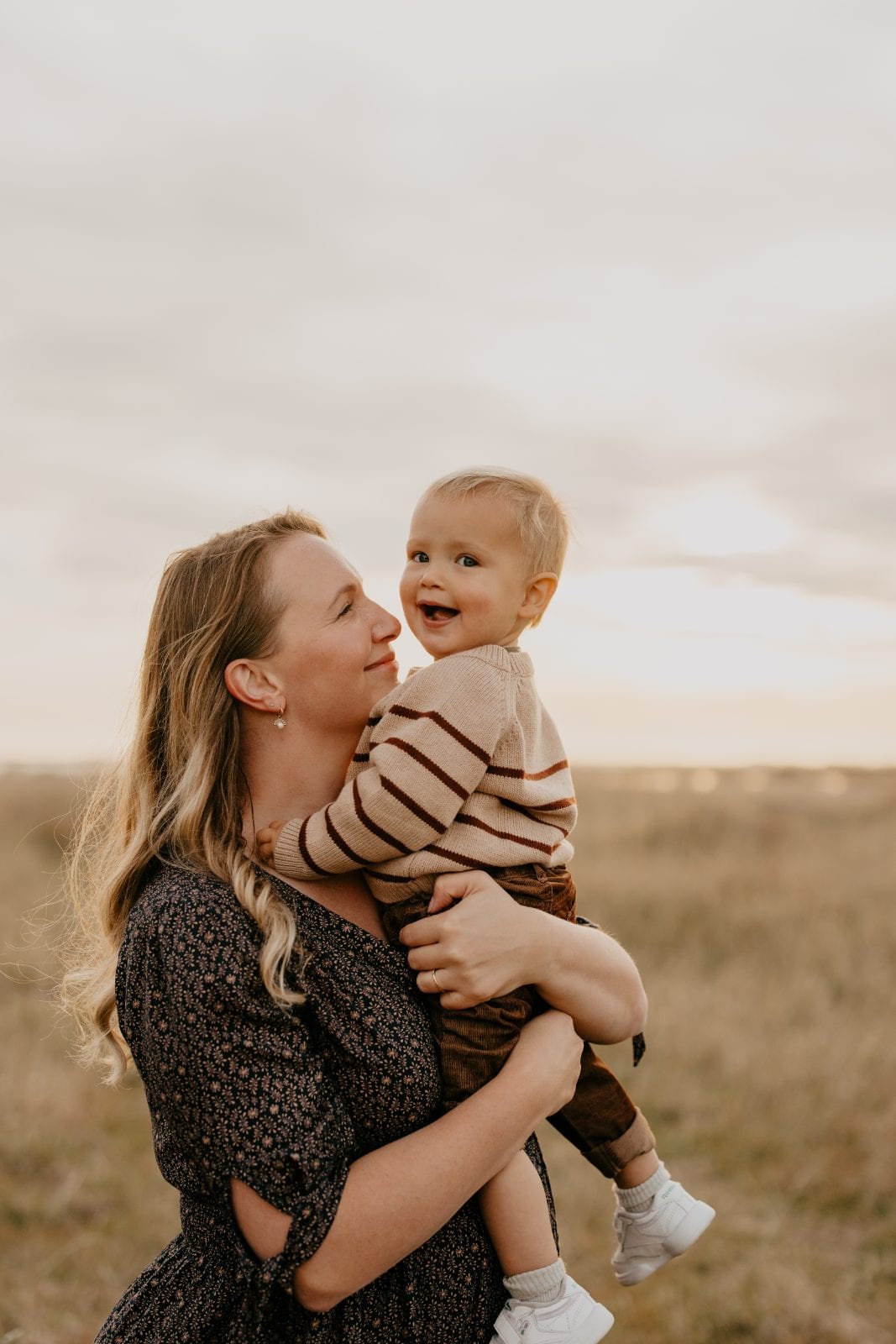 A mother smiles up at her giggling toddler boy as she holds him in a sunlit golden field.