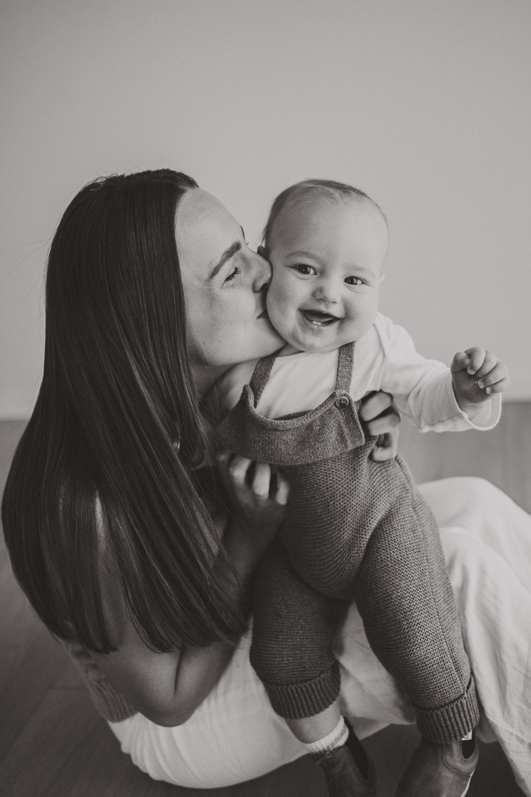 Black and white portrait of a mother kissing her cheerful baby held up in knitted overalls.