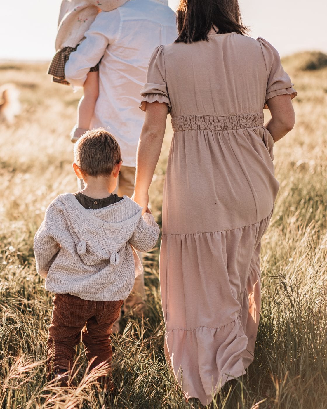 A mother in a blush maxi dress holds her young son's hand as they walk through tall golden grass behind the rest of the family.