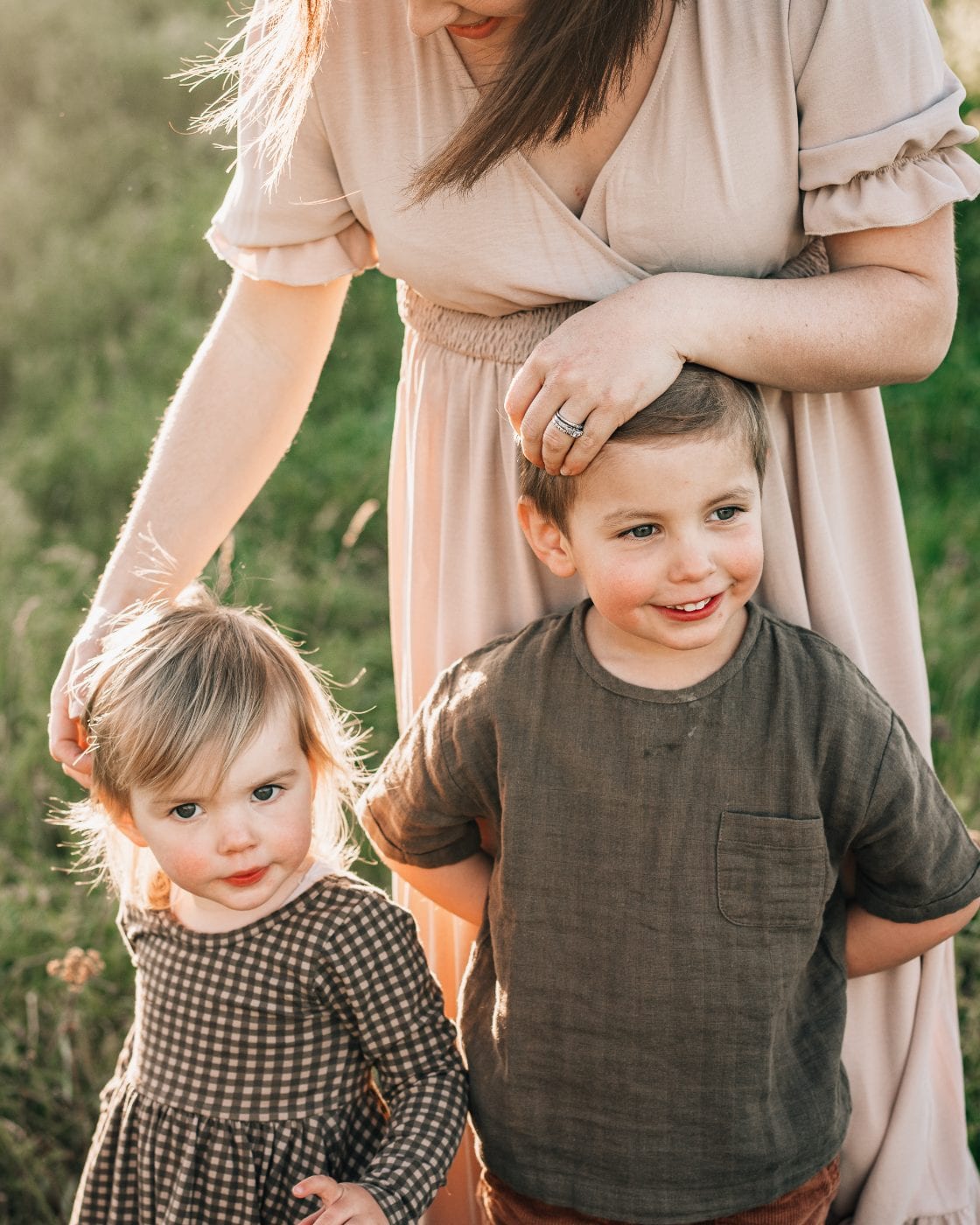 A toddler girl in a gingham dress and her older brother stand with their mother in a sunlit field during a family session.