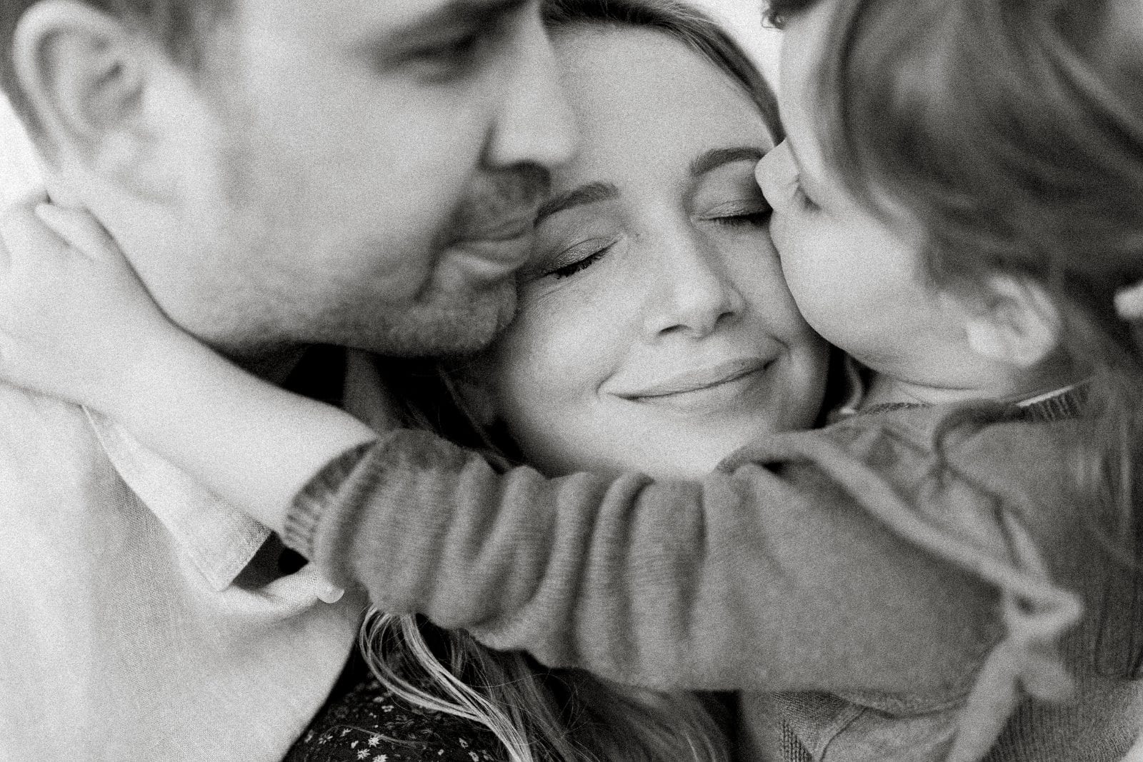 Black and white close-up of parents embracing their young child with gentle cheek kisses.