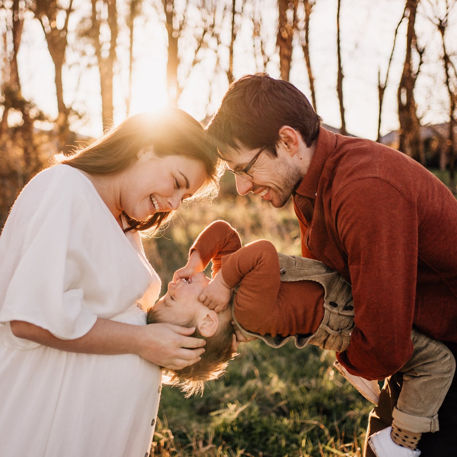 Laughing parents hold their toddler upside down between them in a golden backlit field lined with bare trees.