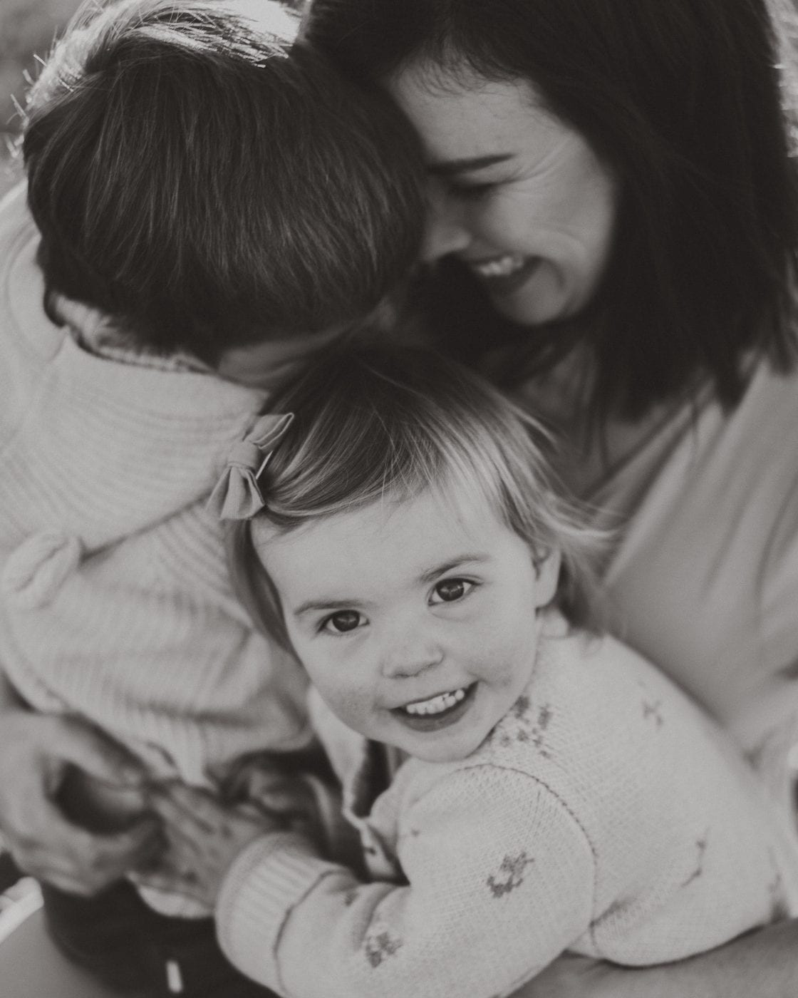 Black and white close-up of parents cuddling their smiling toddler daughter in a soft floral cardigan outdoors.