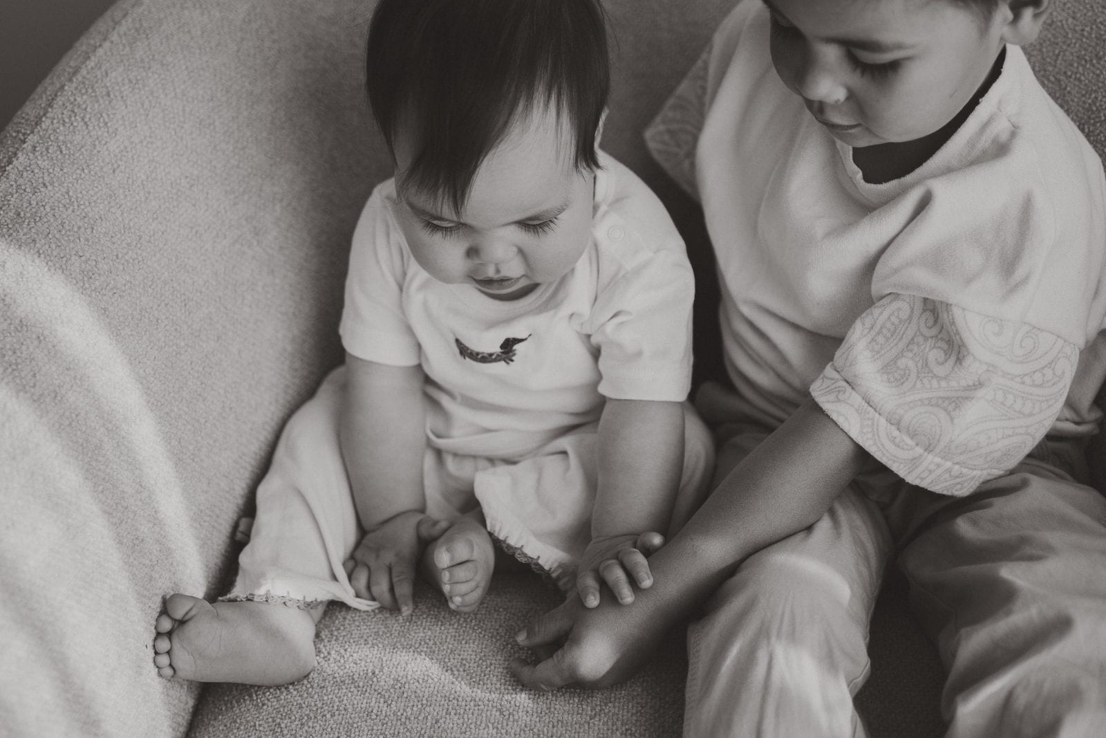 Black and white candid of a baby and older brother sitting close together on a soft armchair.