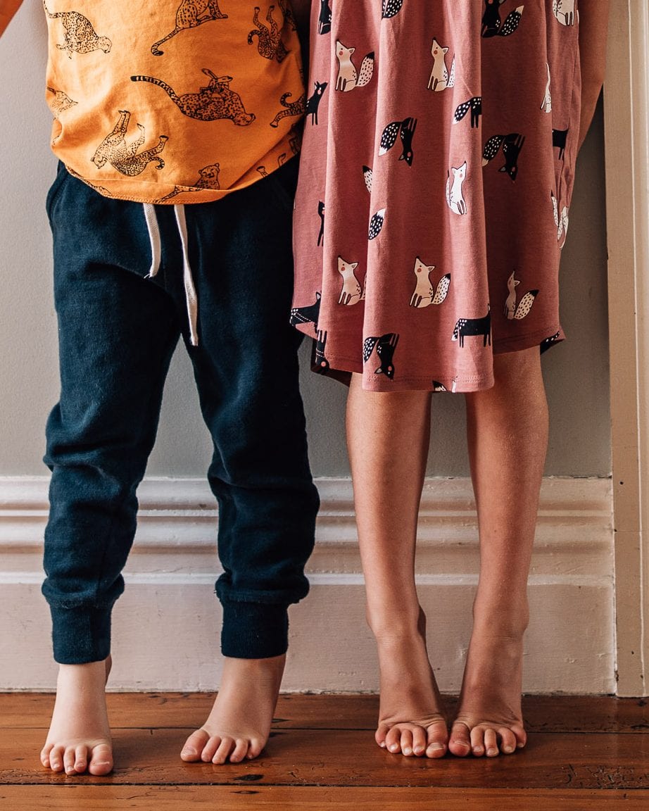 Detail of two young siblings standing barefoot together on a warm wooden floor in patterned outfits.