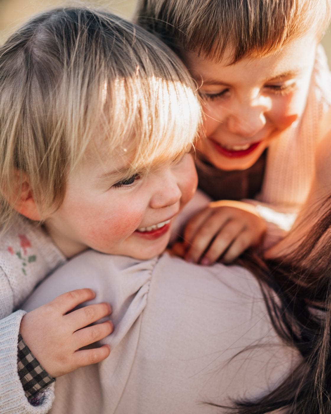 Two young siblings nestle against their mother's shoulder, softly lit by warm late-afternoon sunlight.