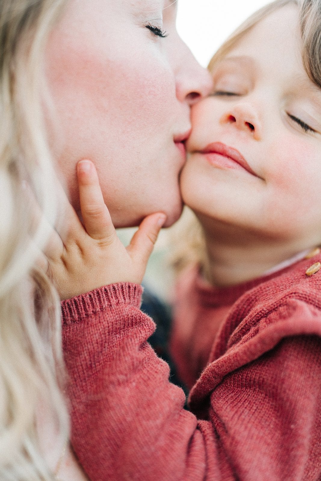 Close-up of two young siblings in rose sweaters as one softly kisses the other's cheek.