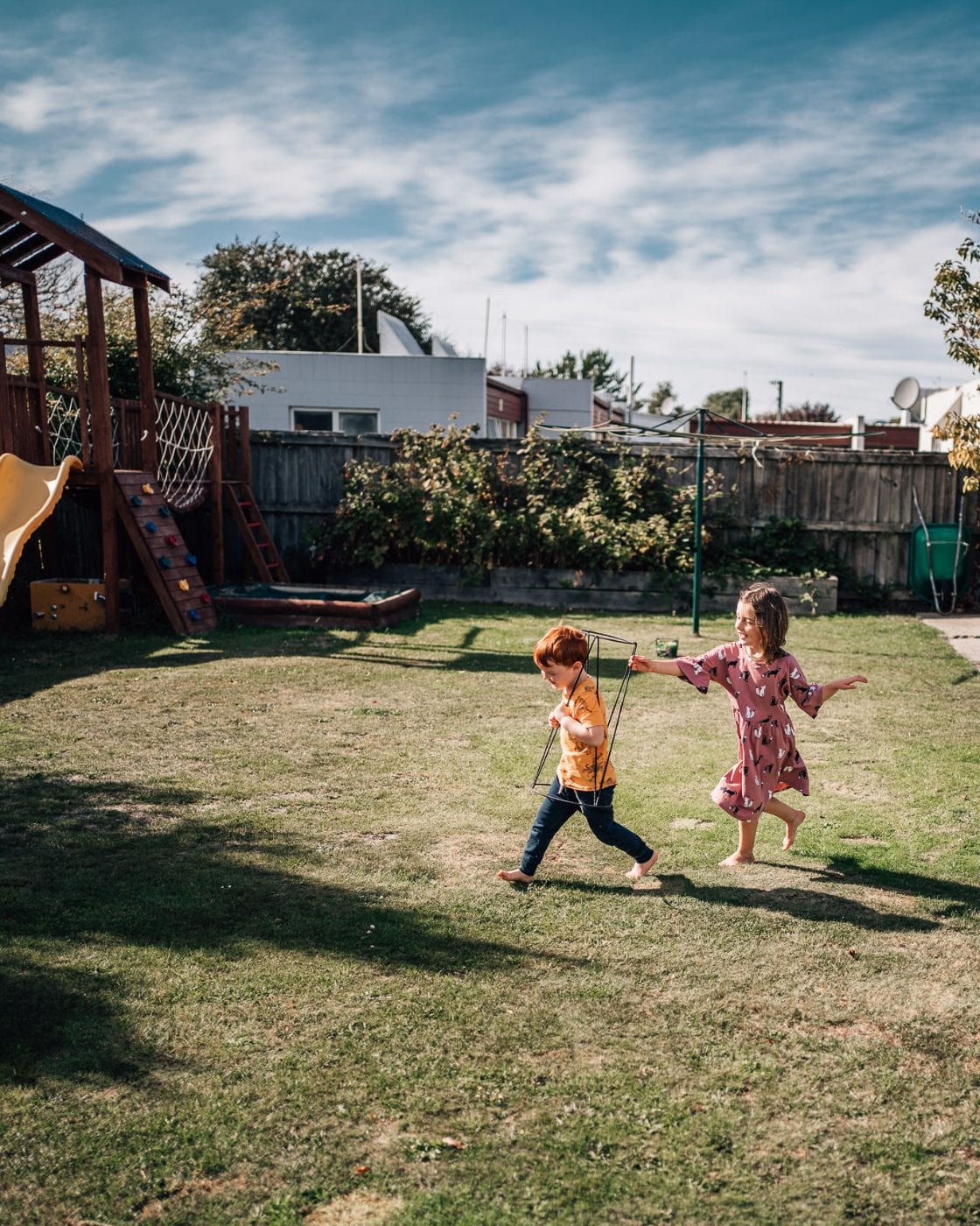 A brother and sister play together barefoot on the grass of a sunny backyard with a play fort.