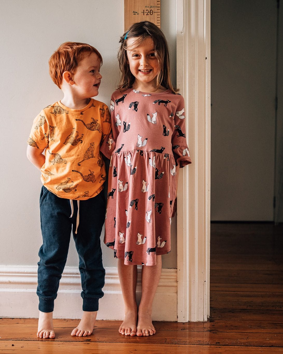 A brother and sister stand side by side against the wall by a wooden height chart, smiling.