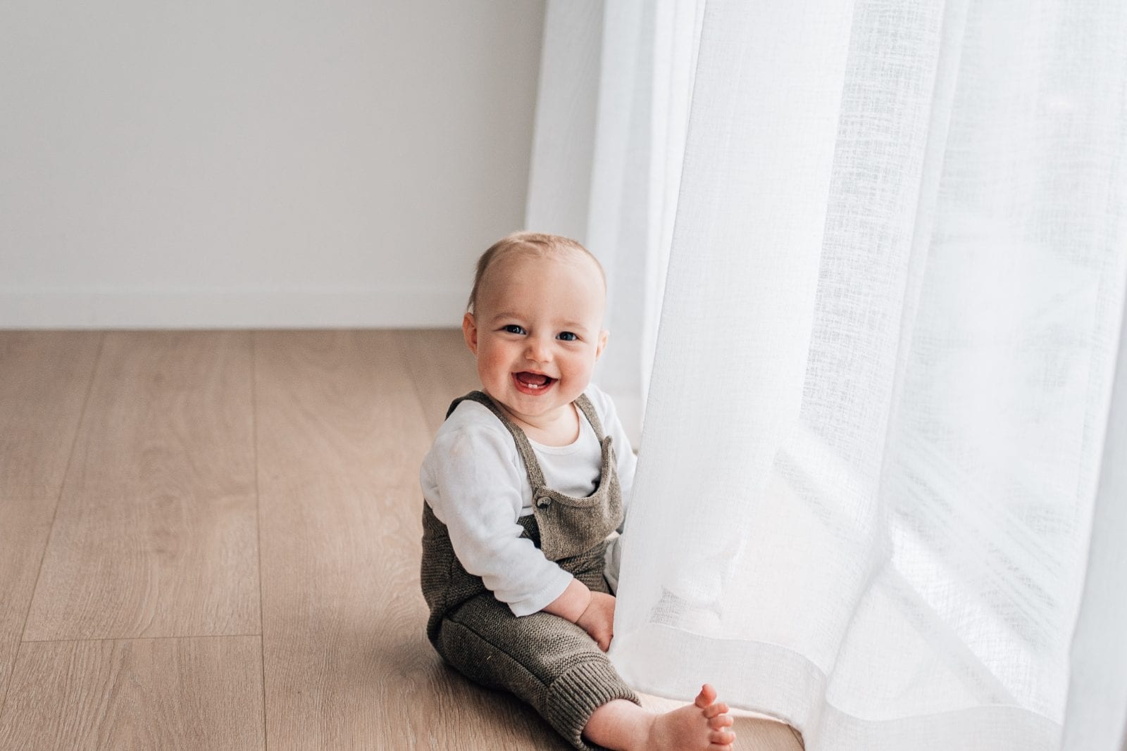 Smiling baby sitting by the studio curtains during a baby milestone session at The Little White Studio in Lincoln