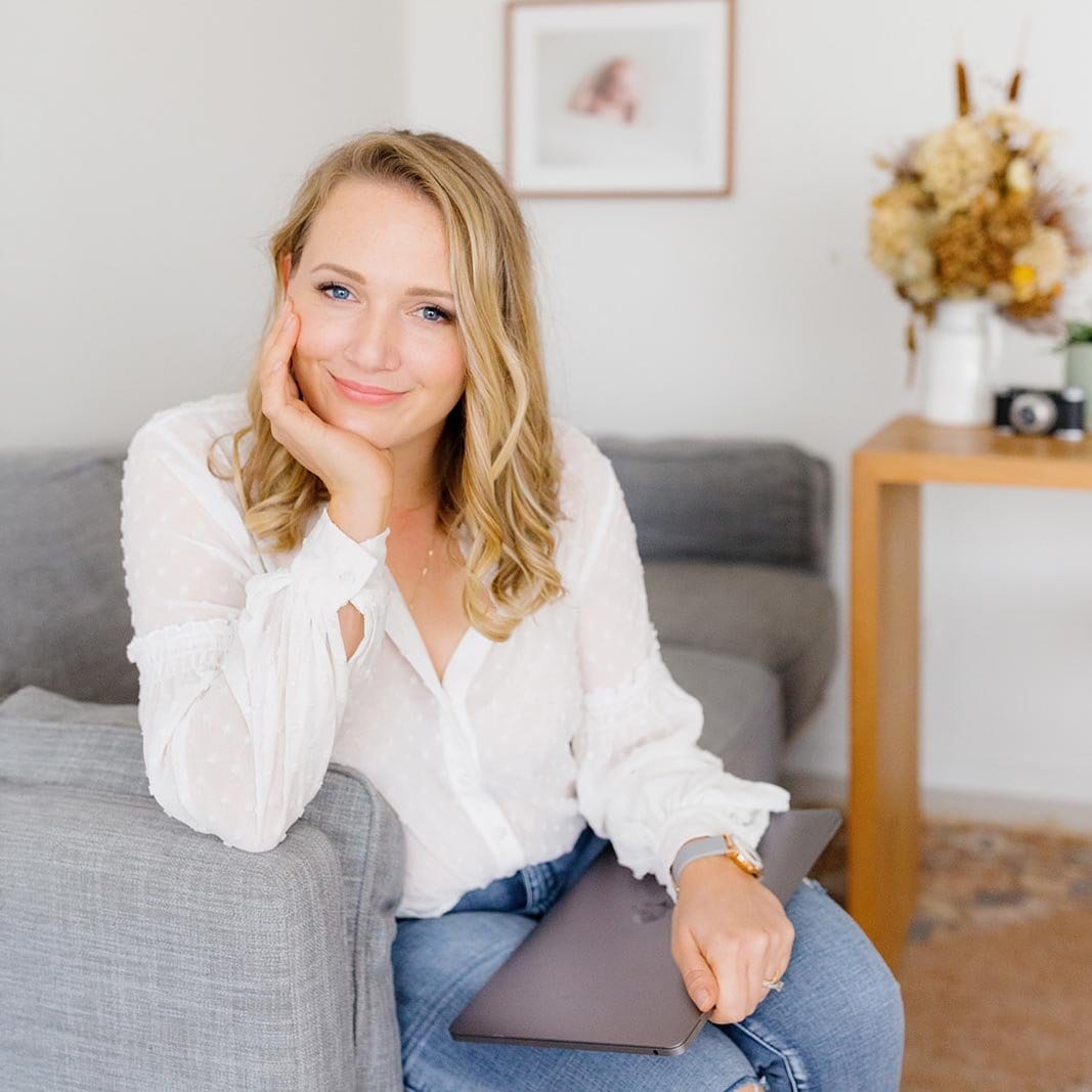 Family photographer Tegan smiles resting her chin on her hand while seated with her laptop in a bright lounge.