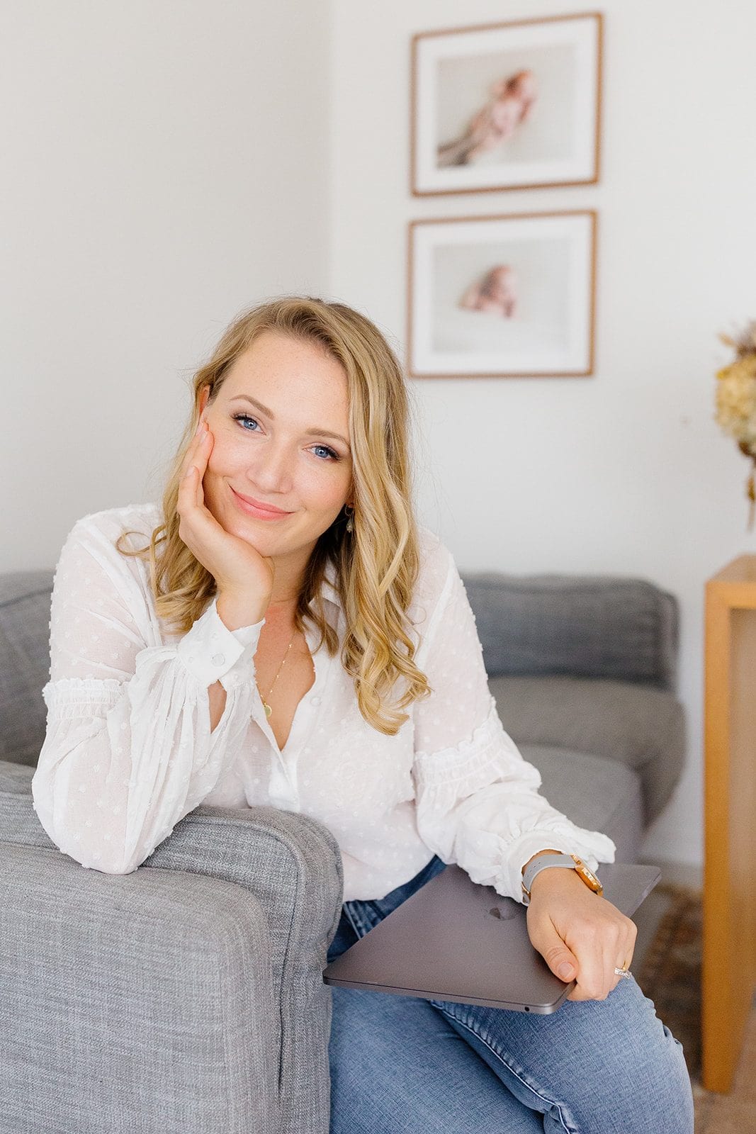 Family photographer Tegan sits on a grey sofa holding her laptop beneath framed newborn prints.