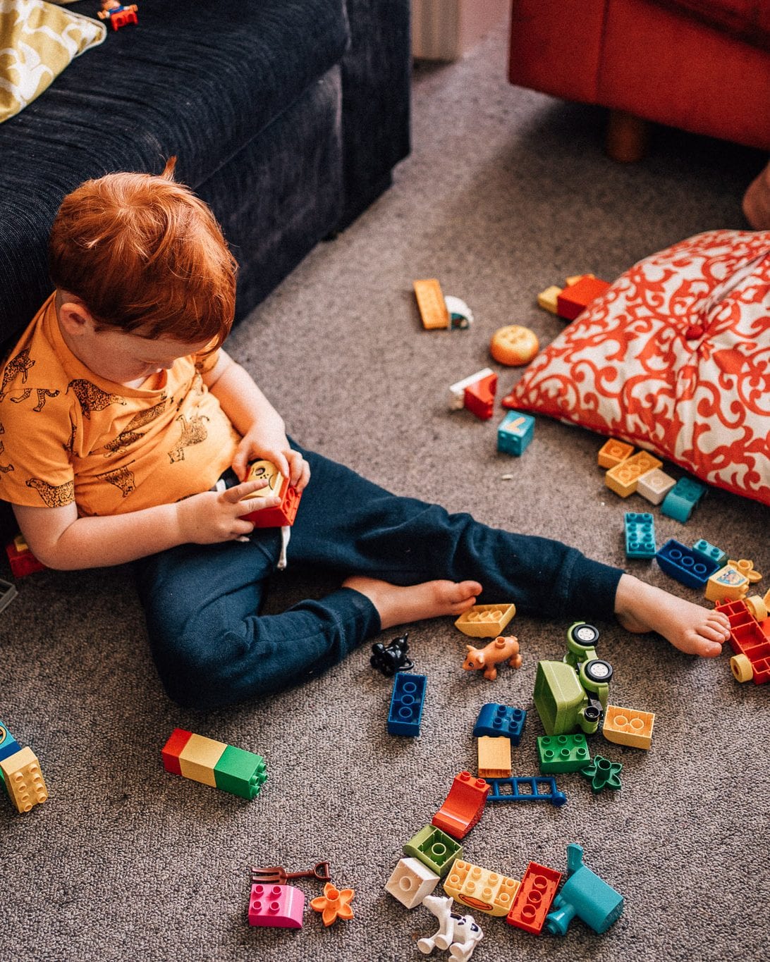 A red-haired toddler boy sits on the floor playing with colourful building blocks in the lounge.