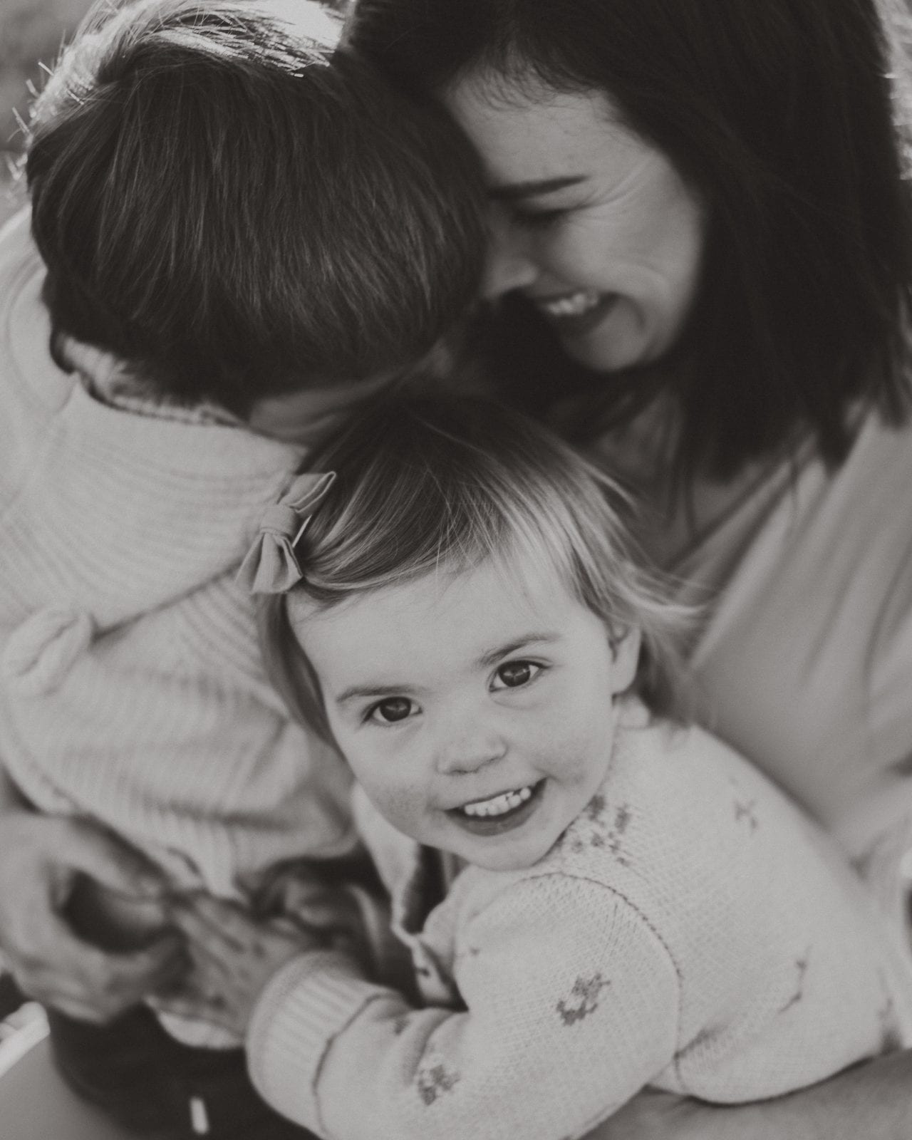Black-and-white close-up of a smiling toddler girl wrapped in a warm cuddle between her two parents.