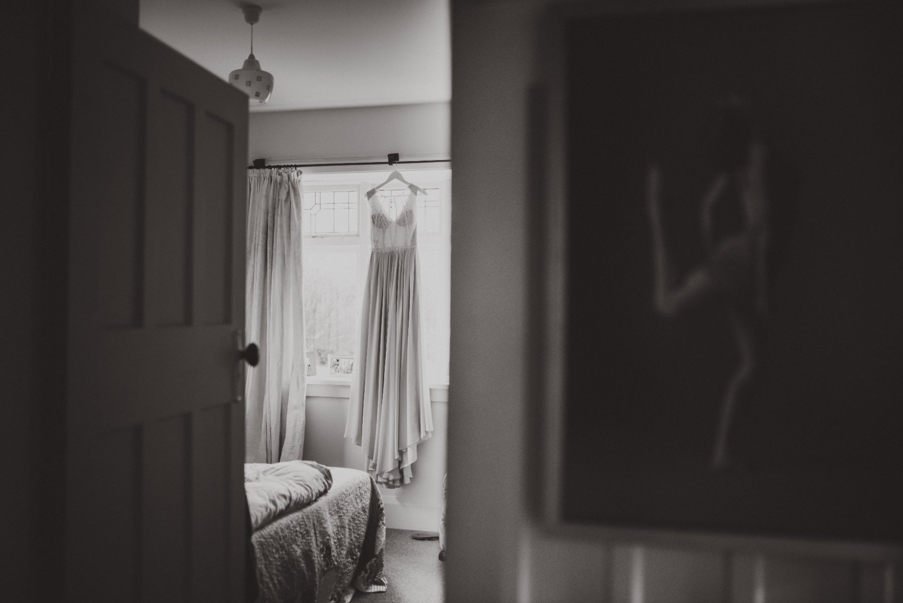 A black and white photo of a wedding dress hanging in a bedroom window, seen through an open doorway