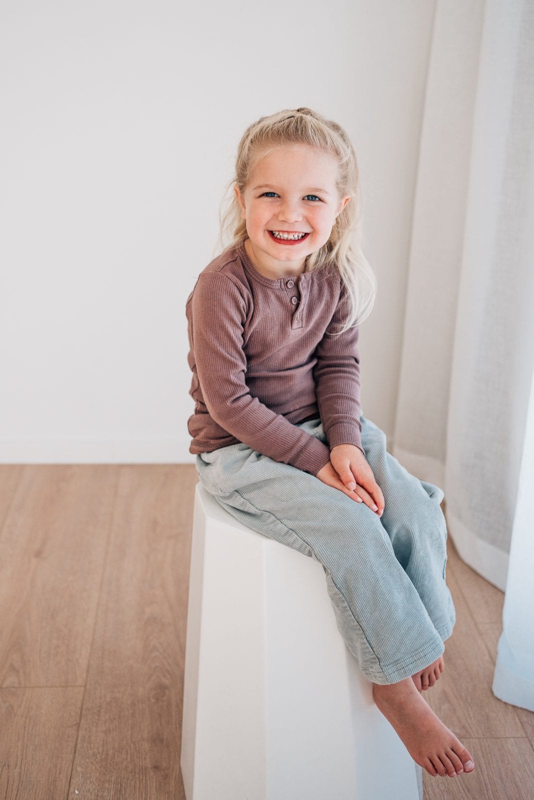 Blonde young girl in a mauve top and cord trousers sitting barefoot on a white plinth, laughing.